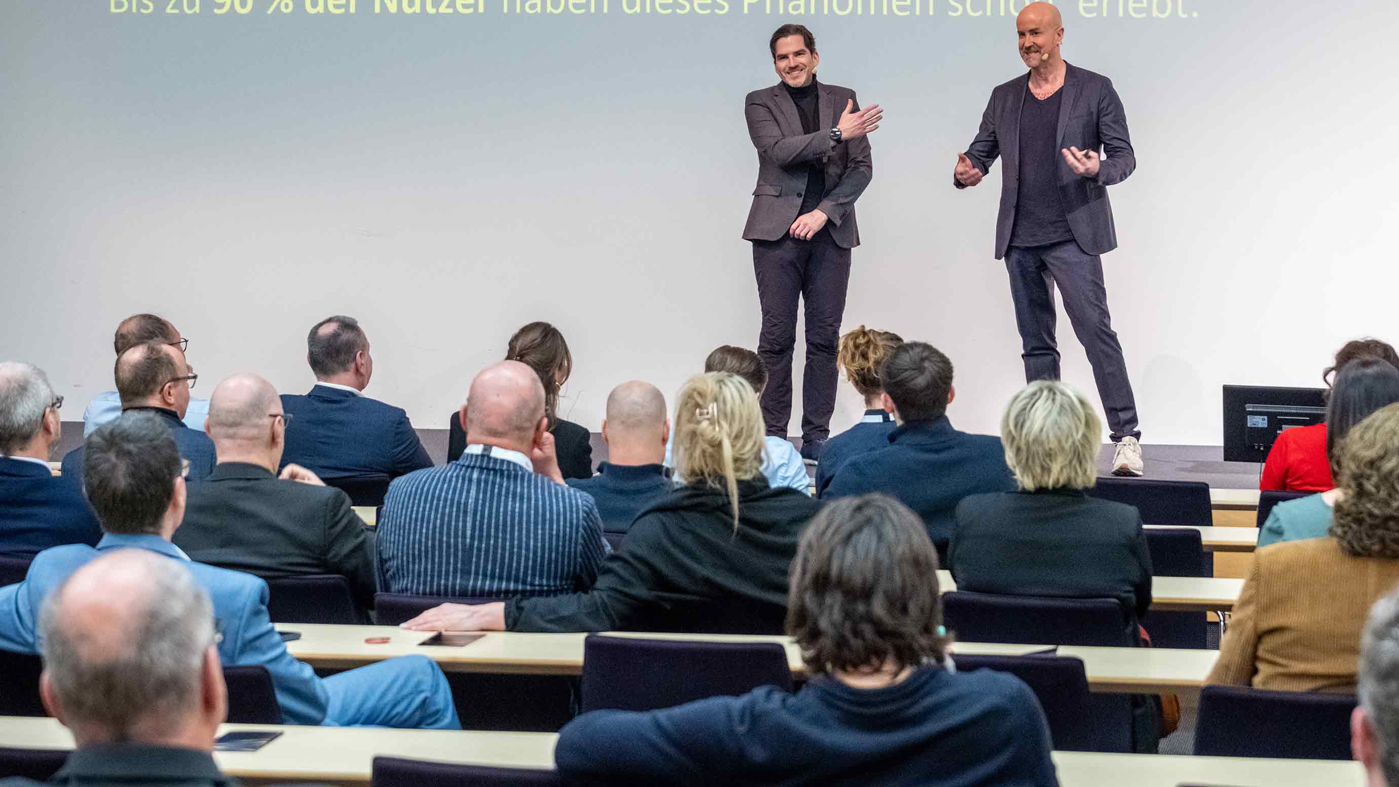 Two speakers standing on a stage and addressing a seated professional audience.