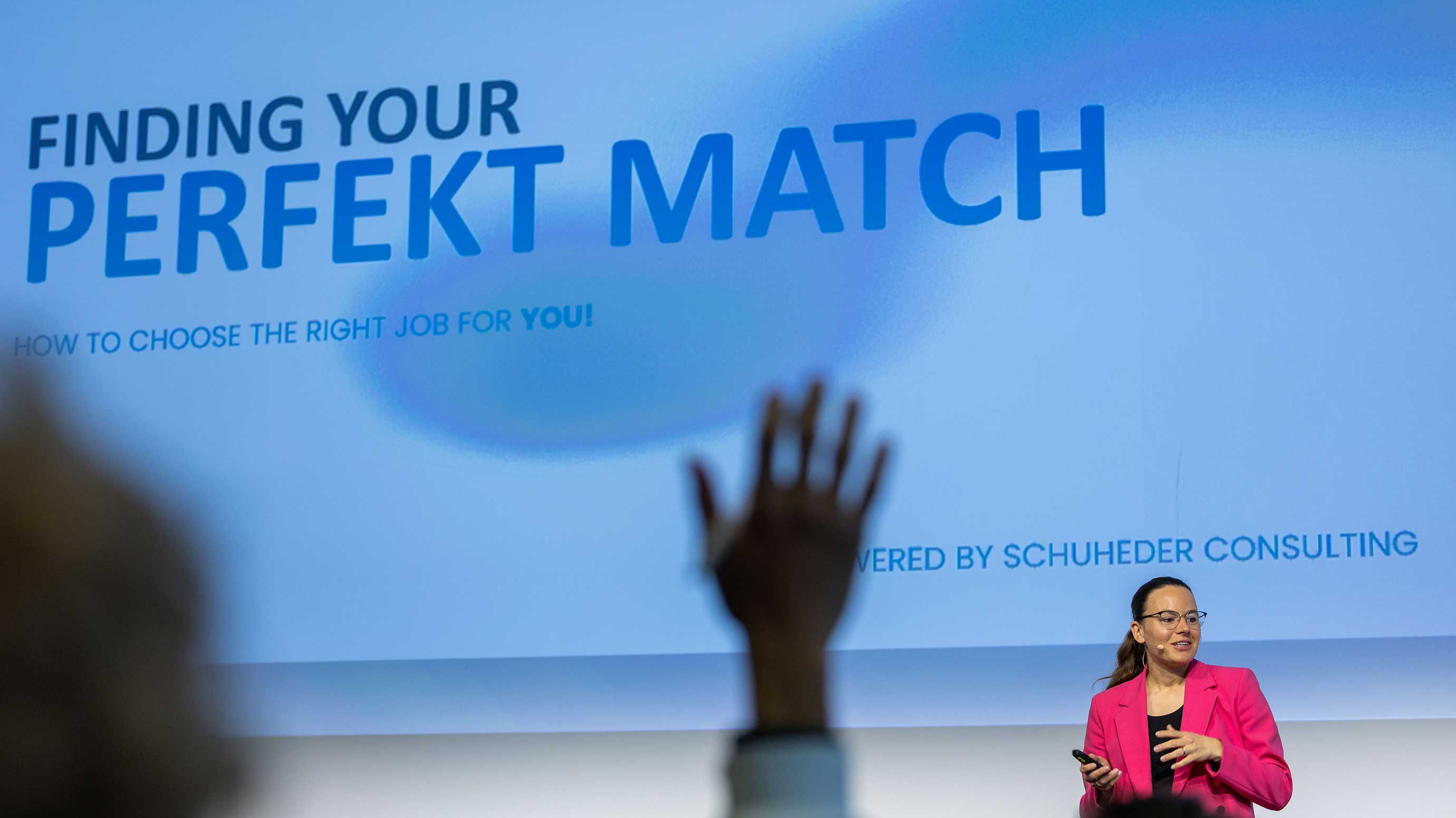 Im Vordergrund eine erhobene Hand, während die Rednerin beim Studententag auf der Bühne steht